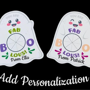 Op de afbeelding: Twee witte spookvormige decoraties met de woorden "YOU ARE FAB BOO LOUS!" en een naam. E&eacute;n heeft groene accenten en zegt "From Ella", de andere heeft paarse accenten en zegt "From Patrick". De zin "Add Personalization" staat onderaan.