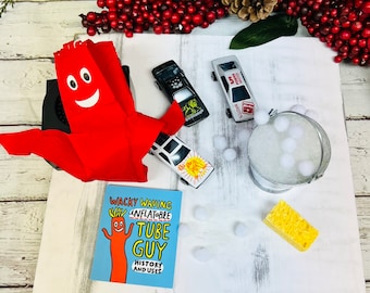 May include: A red inflatable tube man with a smiling face, a blue and white card with the text "Wacky Waving Inflatable Tube Guy History and Uses", a small bucket with white pom-poms, a yellow sponge, and toy cars on a white wooden surface.