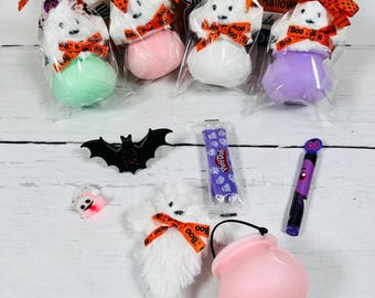 Könnte beinhalten: Eine Sammlung von Halloween-Artikeln, darunter geisterförmige Plüschtiere mit orangefarbenen Schleifen, eine schwarze Fledermausdekoration, ein rosa Kessel und ein lila Stift. Die Geister sind mit bunten Badebomben verpackt.