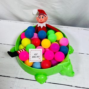 May include: A green turtle-shaped container filled with colorful plastic balls. An elf doll in a red suit and hat sits atop the balls. A small white sign with the text "ELF BALL PIT" is inside the container.