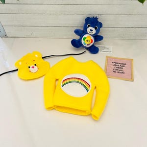 May include: A yellow Care Bear shirt with a rainbow patch and a yellow Care Bear head band. A blue Care Bear plush toy is also in the image. The text on the pink card reads "Spreading Love and Cheer for all to hear!"