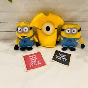 May include: Two yellow plush Minions with blue overalls and a yellow plush Minion shirt with a large eye on the front. The Minions are holding signs that say "One of these things is not like the others!" and "SSSHH! I don't think they notice."
