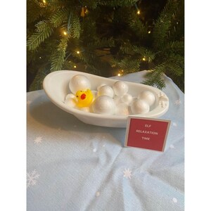 May include: A miniature white bathtub filled with white foam balls and a yellow rubber duck. A small red sign reads "ELF RELAXATION TIME". The scene is set on a blue tablecloth with white snowflake patterns, in front of a Christmas tree.