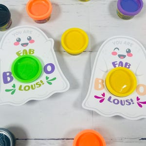 Op de afbeelding: Twee witte spookvormige kaarten met de tekst "YOU ARE FAB BOO LOUS!" en een Play-Doh-container in het midden. Verschillende Play-Doh-containers in oranje, geel, groen, paars en zwart zijn rond de kaarten verspreid.