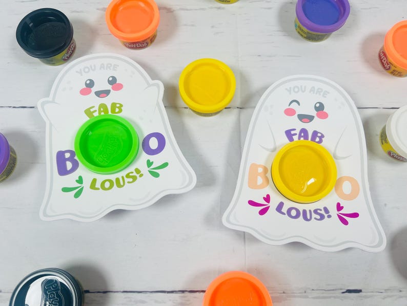 Op de afbeelding: Twee witte spookvormige kaarten met de tekst "YOU ARE FAB BOO LOUS!" en een Play-Doh-container in het midden. Verschillende Play-Doh-containers in oranje, geel, groen, paars en zwart zijn rond de kaarten verspreid.