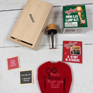 May include: A Christmas Story themed set including a wooden crate marked FRAGILE, a miniature leg lamp, a mini leg lamp kit box, a sticker book, two small square cards with text, and a red sweater with the text You'll Shoot Your eye Out!