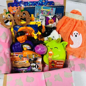 May include: A Halloween-themed gift box filled with various items. Includes pumpkin-shaped decorations, a plush green monster, a tutu, books, bracelets, and candy. The box is lined with pink paper featuring ghost patterns.