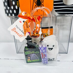 Puede incluir: Una bolsa de regalo transparente llena de golosinas de Halloween. La bolsa incluye un mini rompecabezas, Play-Doh y un fantasma de peluche. Una etiqueta con el texto "You've Been Booed!" est&aacute; adjunta con una cinta naranja.