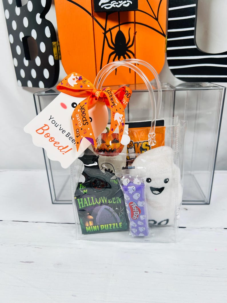 Puede incluir: Una bolsa de regalo transparente llena de golosinas de Halloween. La bolsa incluye un mini rompecabezas, Play-Doh y un fantasma de peluche. Una etiqueta con el texto "You've Been Booed!" est&aacute; adjunta con una cinta naranja.