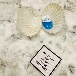 May include: A white seashell with a blue swirl inside. The seashell is open and a blue glass stone is inside. The text on the white sign reads "What can I say, except you're welcome?"