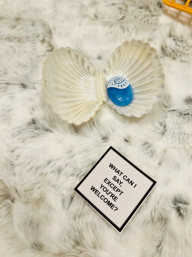 May include: A white seashell with a blue swirl inside. The seashell is open and a blue glass stone is inside. The text on the white sign reads "What can I say, except you're welcome?"