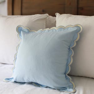Op de afbeelding: Een lichtblauw sierkussen met een geschulpte rand en een witte rand. Het vierkante kussen ligt op een wit bed met twee andere witte kussens op de achtergrond. Het hoofdbord is van hout.