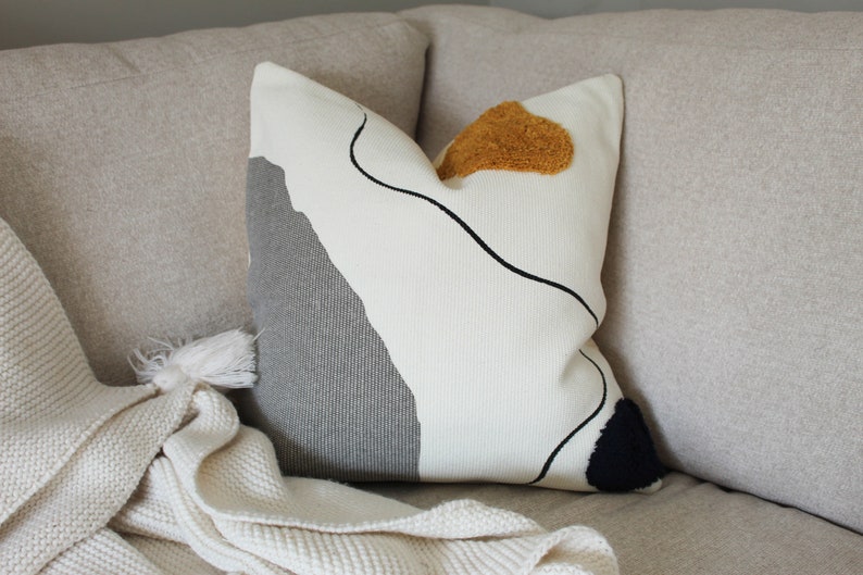Op de afbeelding: Een wit kussen met een abstract ontwerp in grijs, zwart en geel. Het kussen heeft een textuur en is versierd met een zwarte lijntekening en een pluche gele patch.