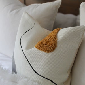 Op de afbeelding: Een wit kussen met een zwart abstract ontwerp en een geel, pluche accent. Het kussen ligt op een wit bed met andere witte kussens.