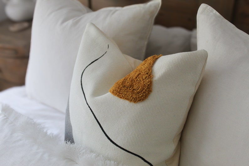 Op de afbeelding: Een wit kussen met een zwart abstract ontwerp en een geel, pluche accent. Het kussen ligt op een wit bed met andere witte kussens.