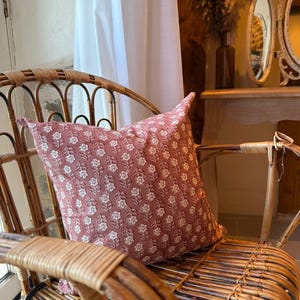 May include: A decorative pillow with a floral pattern in shades of dusty rose and white rests on a wicker chair. The pillow's design features a repeating pattern of stylized flowers. The chair is made of woven rattan.