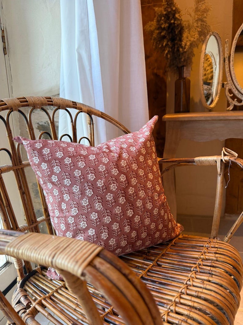 May include: A decorative pillow with a floral pattern in shades of dusty rose and white rests on a wicker chair. The pillow's design features a repeating pattern of stylized flowers. The chair is made of woven rattan.
