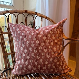 May include: A square decorative pillow with a floral pattern in shades of dusty rose and cream. The pillow is placed on a woven rattan chair, creating a cozy and inviting aesthetic. The pillow's design features a repeating pattern of stylized flowers.