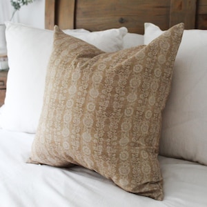 以下が含まれることがあります： ベージュの花柄のクッション。正方形のクッションは白いベッドの上に置かれています。柔らかく、質感のある外観で、寝室の装飾にエレガントさを添えています。