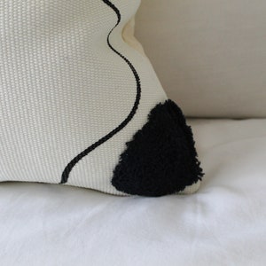 Op de afbeelding: Wit kussen met een zwart abstract ontwerp en een zwarte, pluche patch.