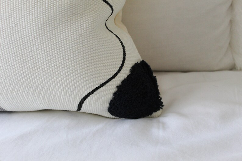 Op de afbeelding: Wit kussen met een zwart abstract ontwerp en een zwarte, pluche patch.