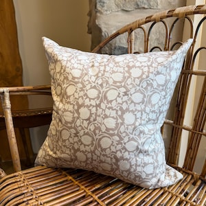 May include: Square cushion with a cream and taupe floral pattern. The cushion is placed on a wicker chair, highlighting its design and texture. The fabric features a repeating botanical motif. The cushion is approximately 40cm x 40cm.