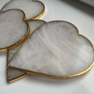 May include: Heart-shaped coasters with a white, marbled appearance and gold-coloured edges. The coasters are made of a stone-like material and are arranged in a slightly overlapping pattern. The gold trim adds a touch of elegance to the design.