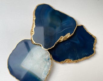 May include: Three irregular, polished agate slices in shades of deep navy blue and translucent white. Each stone coaster features a natural, jagged edge finished with a metallic gold trim, showcasing unique concentric banding and crystalline textures.