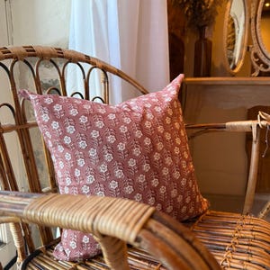 May include: A dusty rose pillow with a white floral pattern rests on a woven rattan chair. The pillow's design features a repeating pattern of stylized flowers. The chair's natural wood tones complement the pillow's color, creating a cozy, inviting aesthetic.