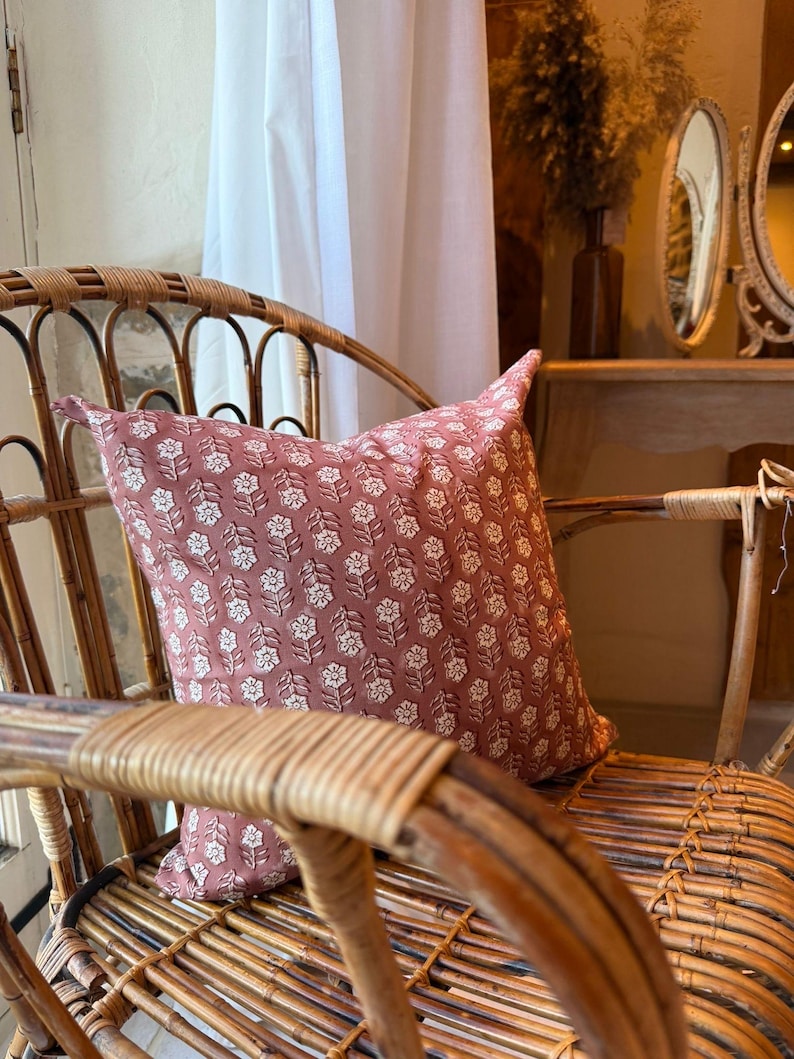 May include: A dusty rose pillow with a white floral pattern rests on a woven rattan chair. The pillow's design features a repeating pattern of stylized flowers. The chair's natural wood tones complement the pillow's color, creating a cozy, inviting aesthetic.