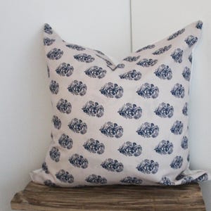 Puede incluir: Un cojín cuadrado con un fondo beige claro y un patrón repetido de diseños florales azul oscuro. El cojín está sobre una superficie de madera y contra una pared blanca.