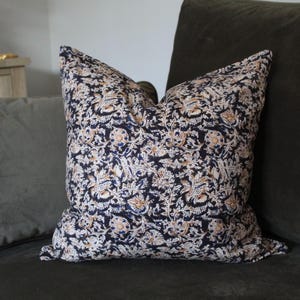 May include: A square throw pillow featuring a dense, intricate floral pattern in shades of dark navy blue, cream, and muted orange. The fabric displays a repeating botanical motif with stylized leaves and blossoms covering the entire surface.