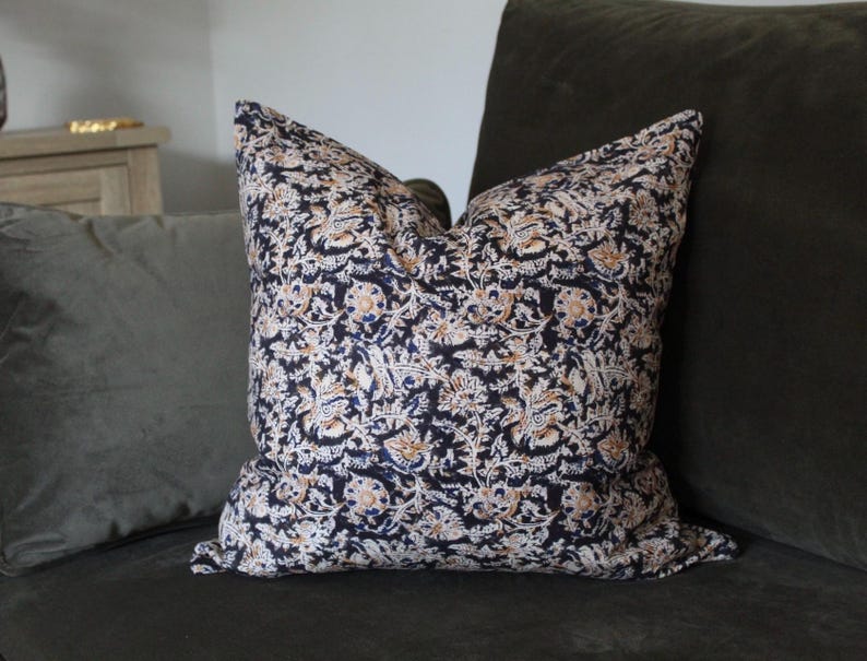 May include: A square throw pillow featuring a dense, intricate floral pattern in shades of dark navy blue, cream, and muted orange. The fabric displays a repeating botanical motif with stylized leaves and blossoms covering the entire surface.