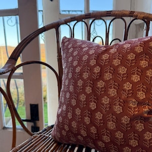 May include: A decorative pillow with a floral pattern in shades of burgundy and cream rests on a woven rattan chair. The pillow's design features repeating stylized flower and leaf motifs, adding a touch of elegance to the seating arrangement.