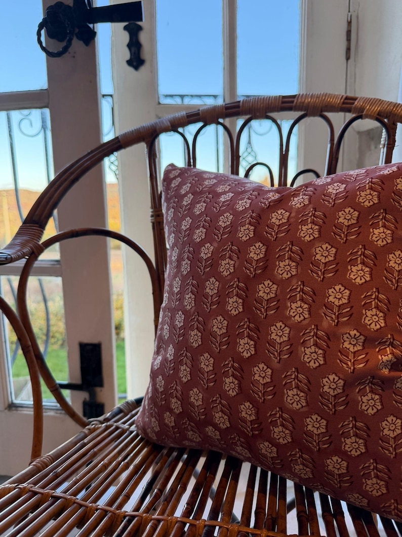 May include: A decorative pillow with a floral pattern in shades of burgundy and cream rests on a woven rattan chair. The pillow's design features repeating stylized flower and leaf motifs, adding a touch of elegance to the seating arrangement.
