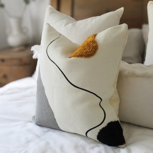 Op de afbeelding: Een wit en grijs kussen met een zwart abstract ontwerp en een gele accent. Het kussen ligt op een wit bed met andere kussen.
