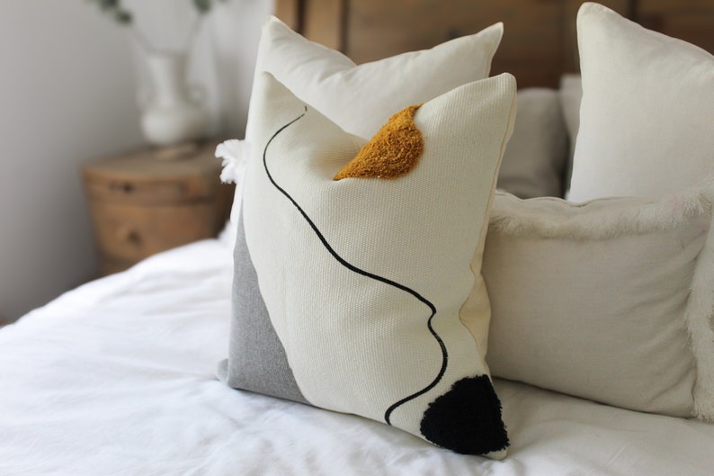 Op de afbeelding: Een wit en grijs kussen met een zwart abstract ontwerp en een gele accent. Het kussen ligt op een wit bed met andere kussen.