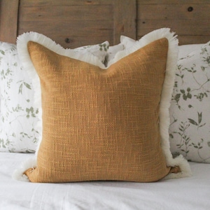 Puede incluir: Una almohada de color mostaza con un borde de flecos blancos. La almohada está sobre una cama blanca con otras almohadas con un patrón floral.