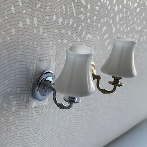May include: A miniature wall sconce with two white lampshades. The sconce has a silver base and a gold-colored arm. The lampshades are bell-shaped and made of a translucent material. The sconce is mounted on a textured, light-colored wall.