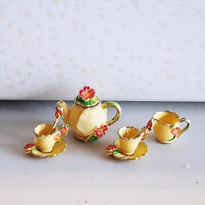 May include: A miniature tea set in cream, featuring a teapot, two teacups with saucers, and a creamer. The set is adorned with red and pink floral designs and green detailing. The teapot is topped with a red flower.