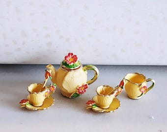 May include: A miniature tea set with a cream-colored teapot, two teacups with saucers, and a creamer. The set is decorated with red and pink flowers and green accents. The teapot has a red flower on top.