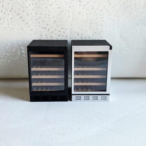 May include: Miniature black and silver wine refrigerators. Each has a glass door revealing wooden shelves. The black refrigerator has a black frame, while the silver refrigerator has a silver frame. Both have a vent at the bottom.