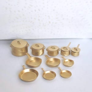 May include: A collection of eleven miniature gold-colored kitchen cookware items, including five lidded pots of varying sizes and six open skillets with handles, all featuring a metallic finish and arranged in a group on a plain surface.