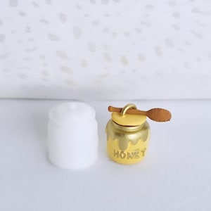 May include: A miniature yellow honey pot charm with a brown wooden dipper attached to the lid and the word HONEY embossed on the front, positioned next to a plain white cylindrical container of similar size and shape.