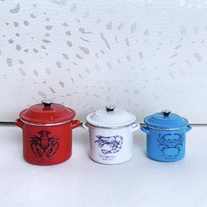 May include: Three miniature enameled pots with lids, featuring a red pot with a lobster graphic, a white pot with a shrimp graphic, and a blue pot with a crab graphic. Each pot is labeled with the text Le Creuset France and has two side handles.
