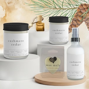 May include: A collection of scented products, including candles, wax melts, and room spray, with the text "cashmere cedar" on the labels. The products are displayed on white pedestals and a wooden platform, with pine needles and a pine cone in the background.