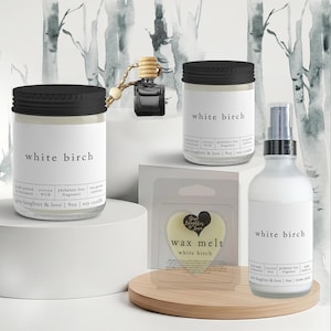 May include: Two glass jars with black lids, a small glass car diffuser, a heart-shaped wax melt in plastic packaging, and a frosted glass spray bottle, all labeled White Birch by Give Laughter & Love. The candles are 9oz and the spray is 4oz.