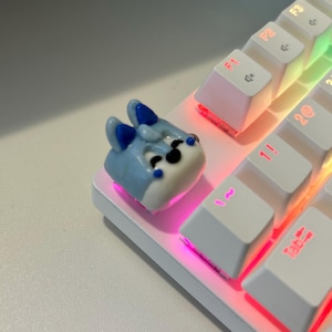 May include: A blue and white character keycap with a smiling face, sitting on a white keyboard with rainbow-colored backlighting.