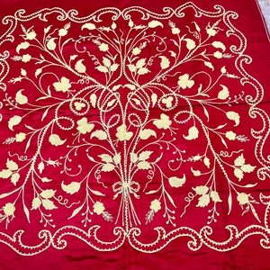 Puede incluir: Un cuadrado de tela roja con un diseño floral bordado en blanco. El diseño presenta un ramo central de flores con hojas y tallos que se extienden hacia afuera. El borde es un diseño de rollo bordado en blanco.