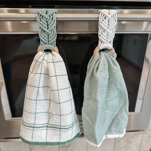 May include: Two green kitchen towels hanging on a silver oven handle. The towels are held in place by macrame loops with wooden rings. One towel has a white and green checkered pattern, and the other has a solid green pattern.